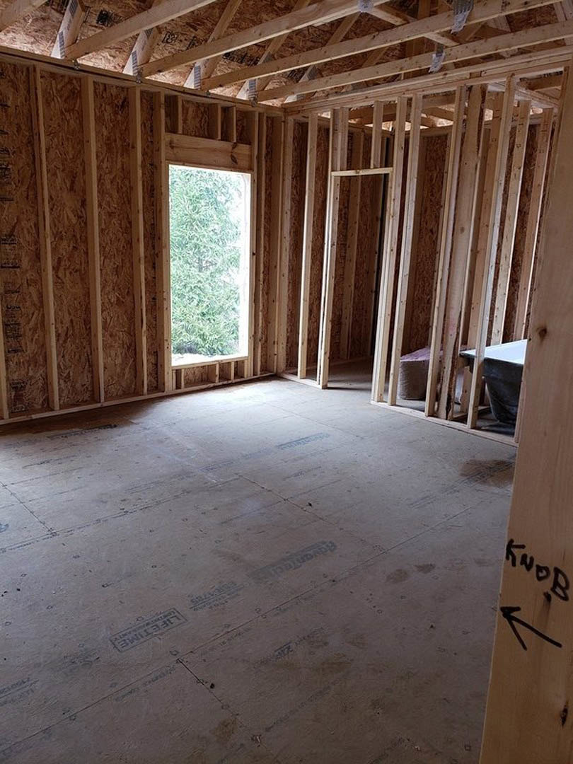 Wood-framed room under construction with exposed beams, unfinished walls, and a window overlooking trees