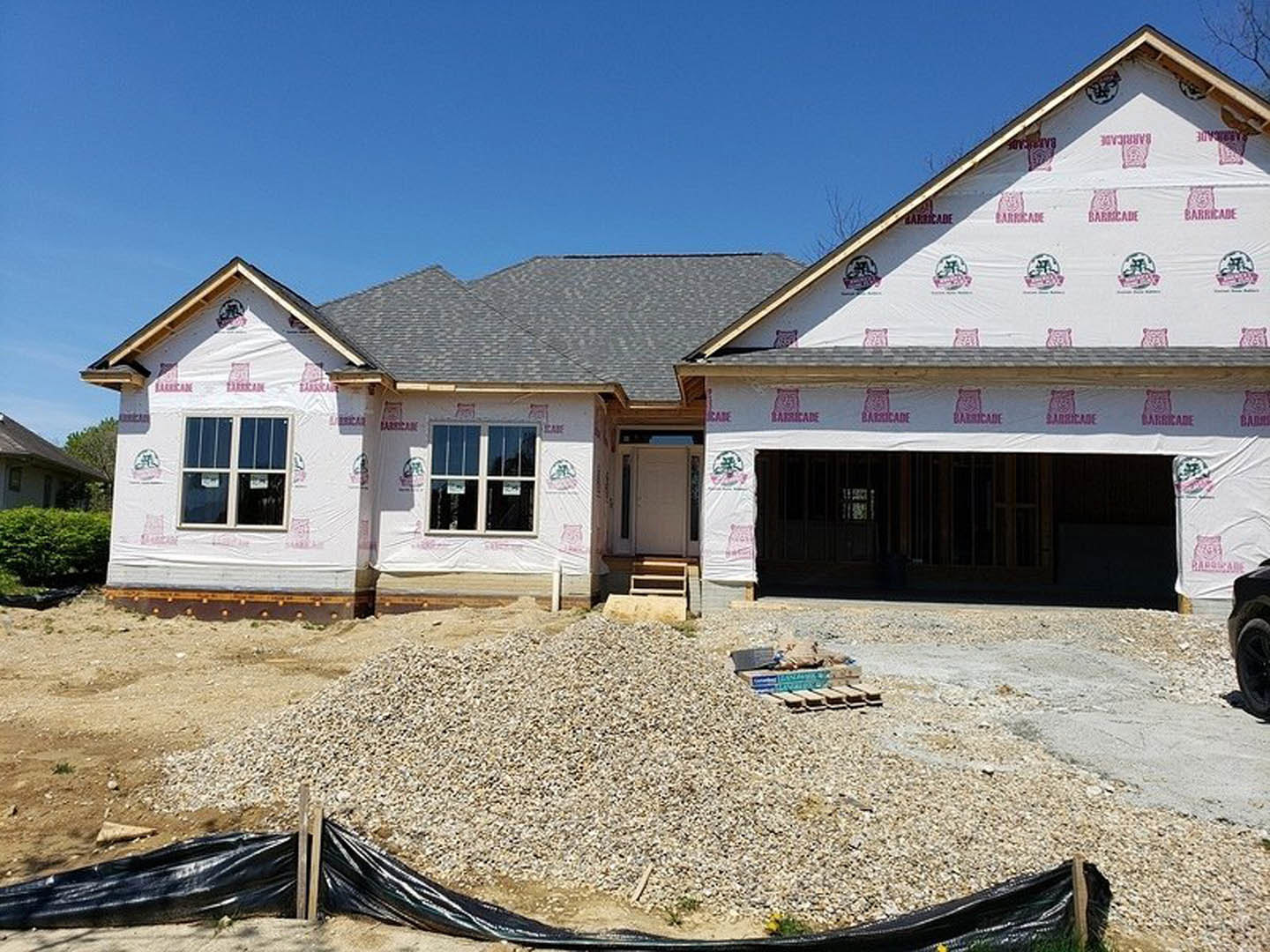 Two-story house under construction with gravel covering the ground, plastic sheeting draped over exterior siding, and unfinished windows visible