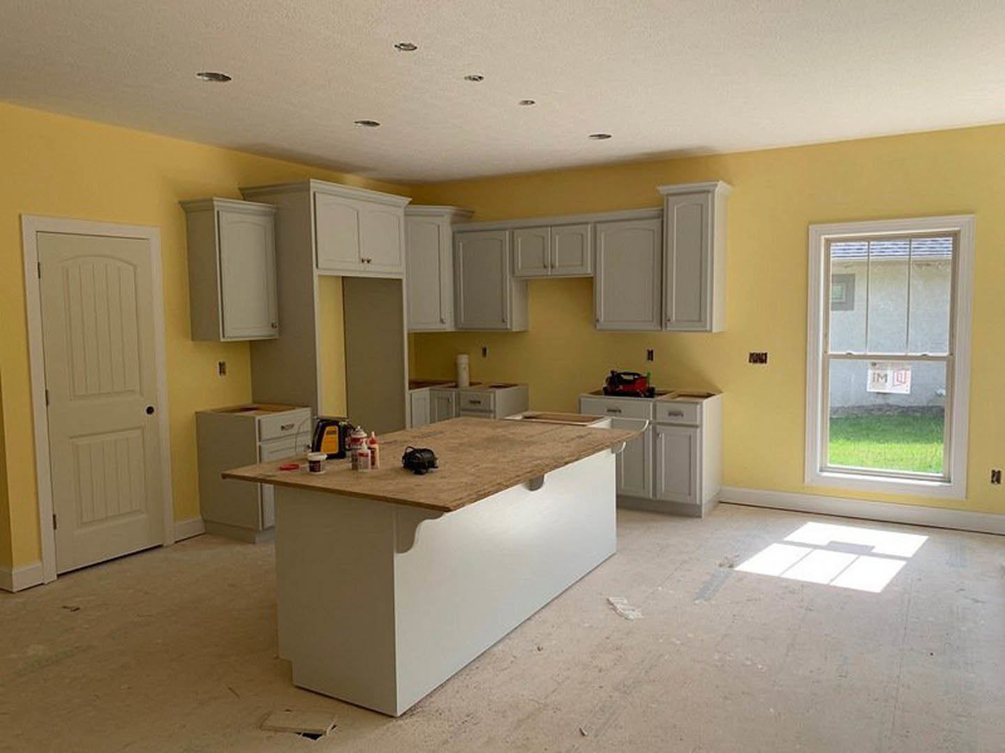 Spacious kitchen featuring a large wood-topped island, white tile floor with shadows, white cabinetry, a window with a sign, and a white door with a black knob.