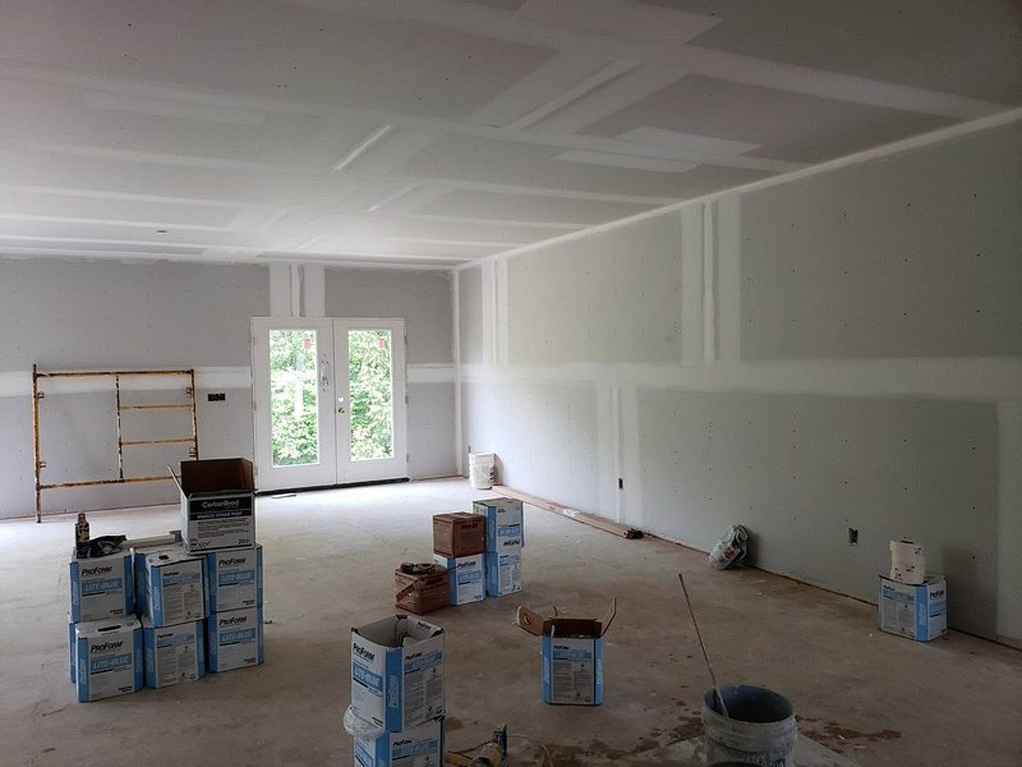 Unfinished room with plaster walls, double glass doors, scattered cardboard boxes and plastic buckets on the floor, exposed rusted metal bars, construction materials visible