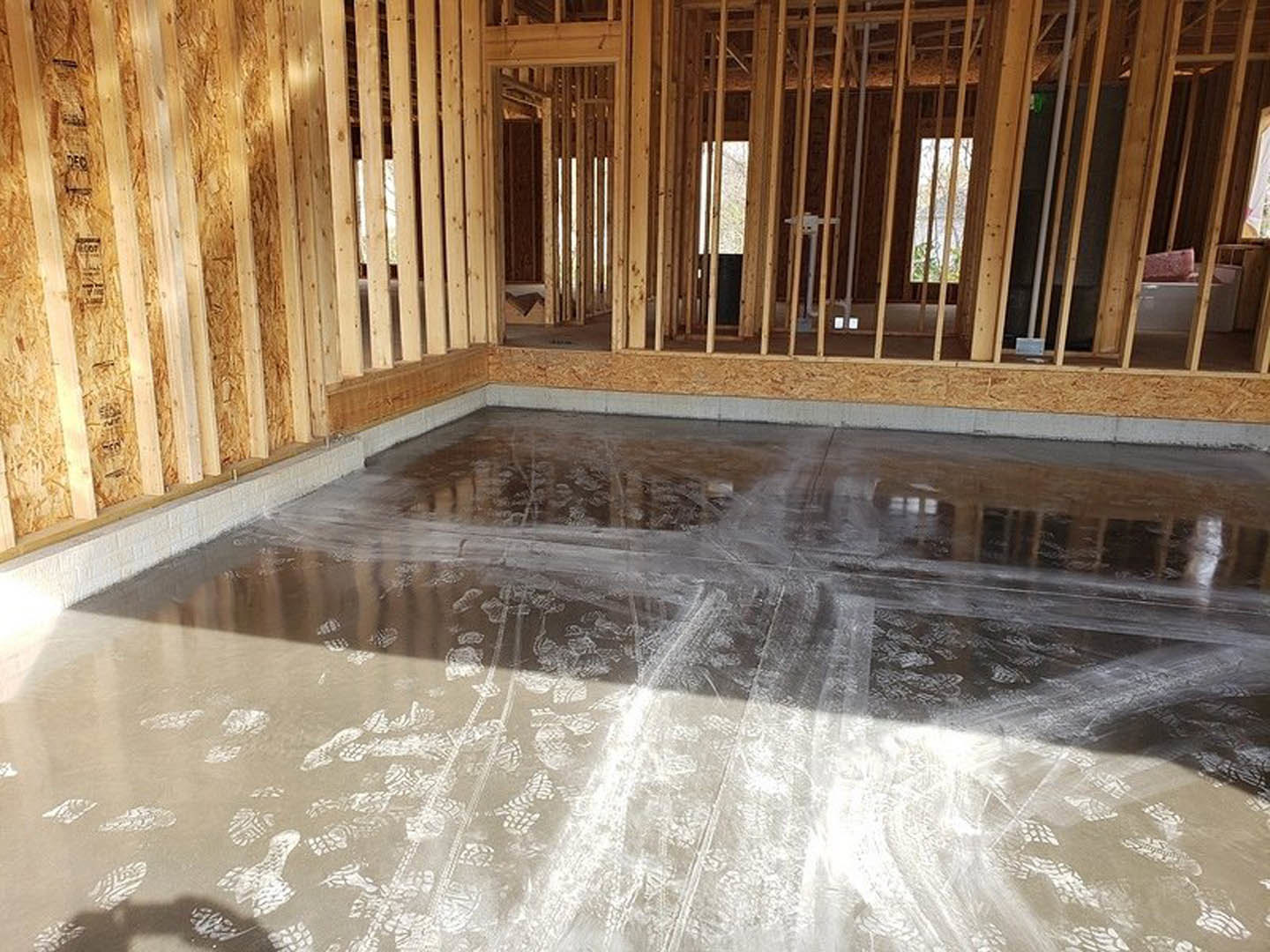 Concrete floor with visible footprints, surrounded by unfinished wooden framing in a residential interior under construction.