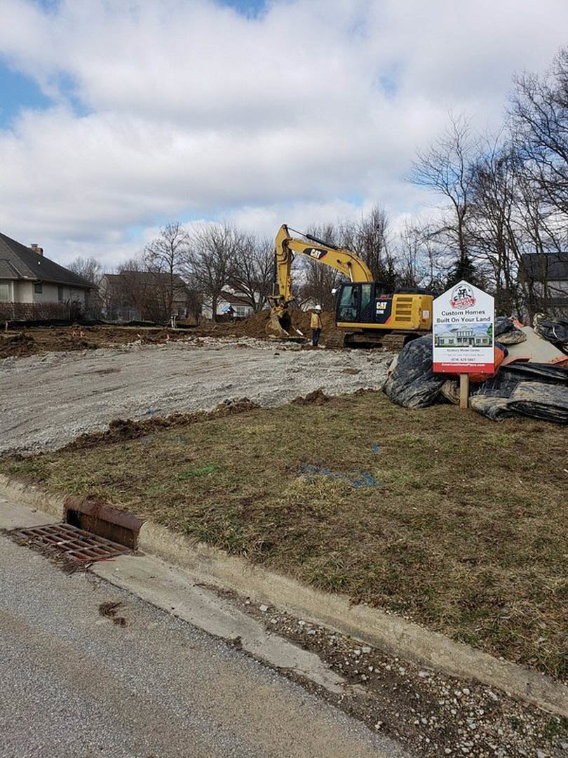Bulldozer parked on dirt construction site near trees, metal grate on ground, fenced area with house sign in background, man standing beside yellow construction vehicle