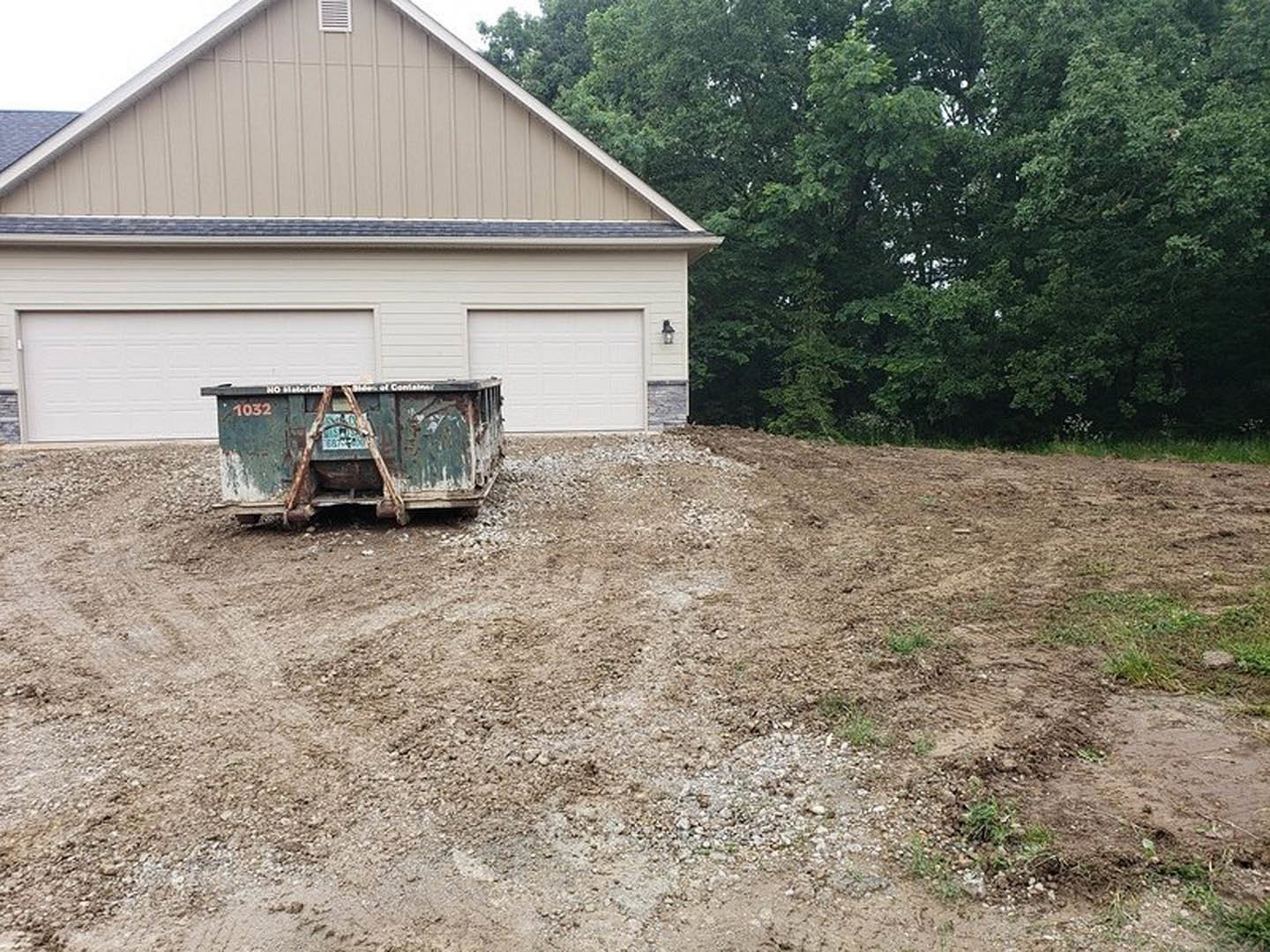 Green dumpster on dirt lot in front of partially constructed house with trees in background