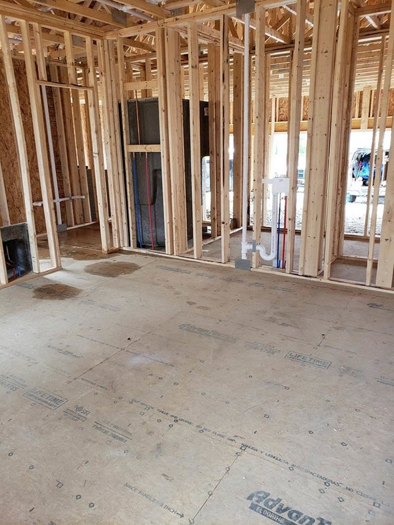 Wood-framed room under construction with exposed beams, wall insulation, visible plumbing pipes, and unfinished floor