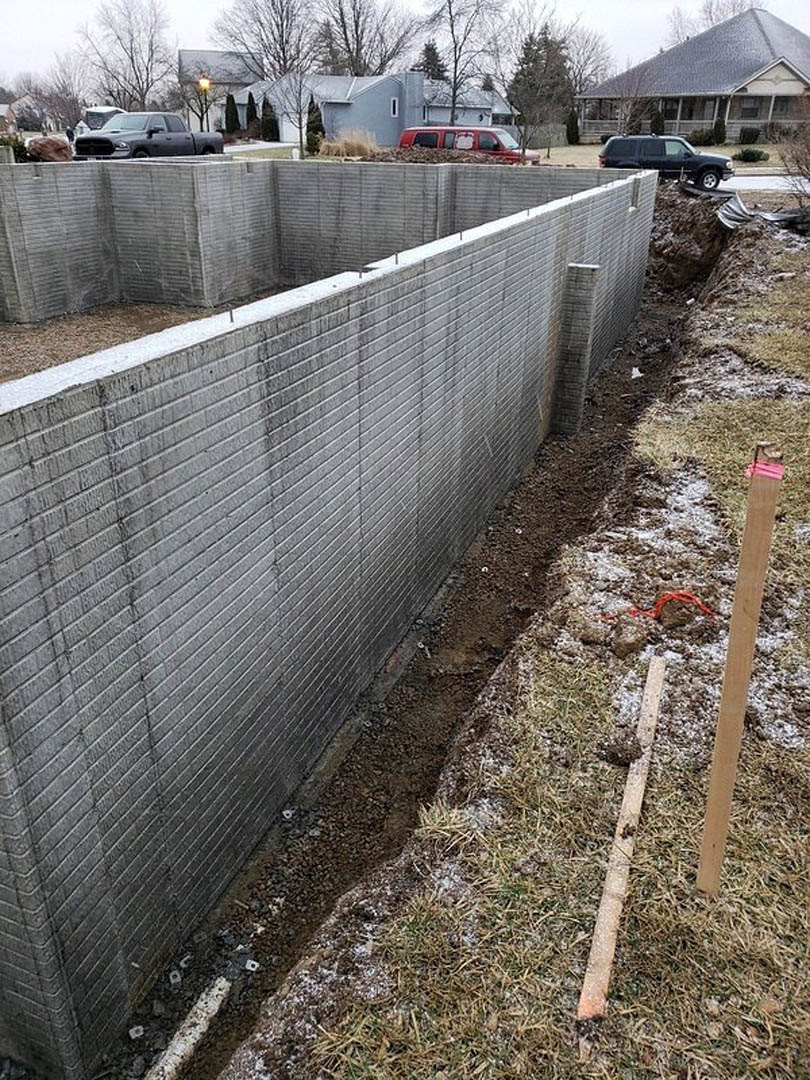 Concrete retaining wall with circular opening in the ground, brown wooden post set in grassy area, house with pitched roof and car parked on street in background, bare trees and