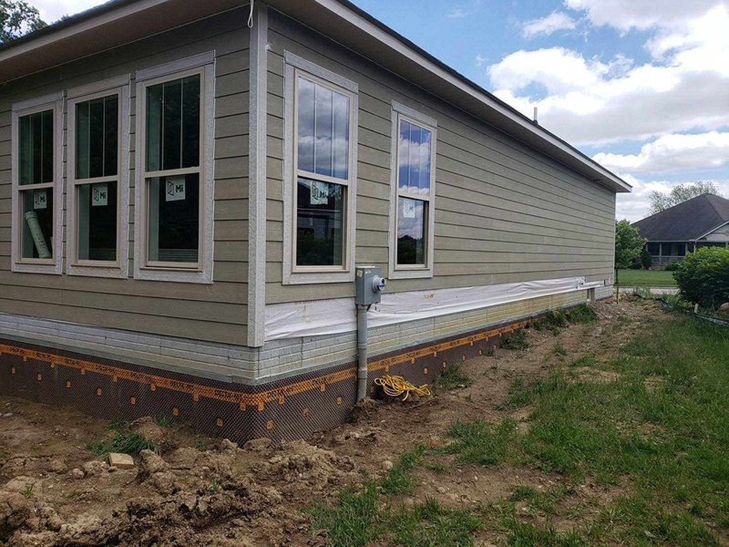 Partially built house with black roof, exposed foundation, white siding, front porch, grassy yard, and blue sky with scattered clouds