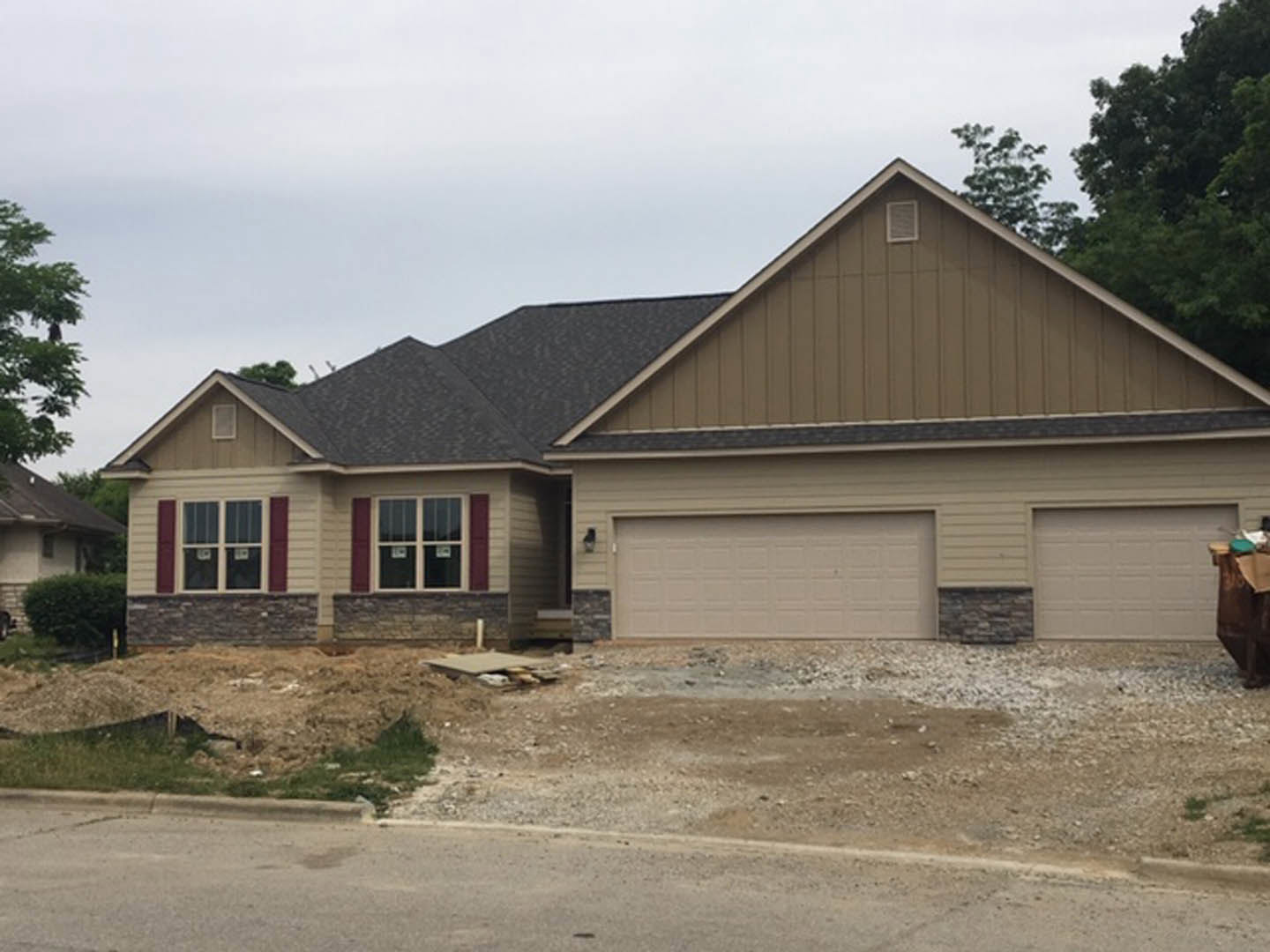 Two-story house under construction with exposed framing, attached garage, dirt hill in foreground, and mature trees surrounding the property