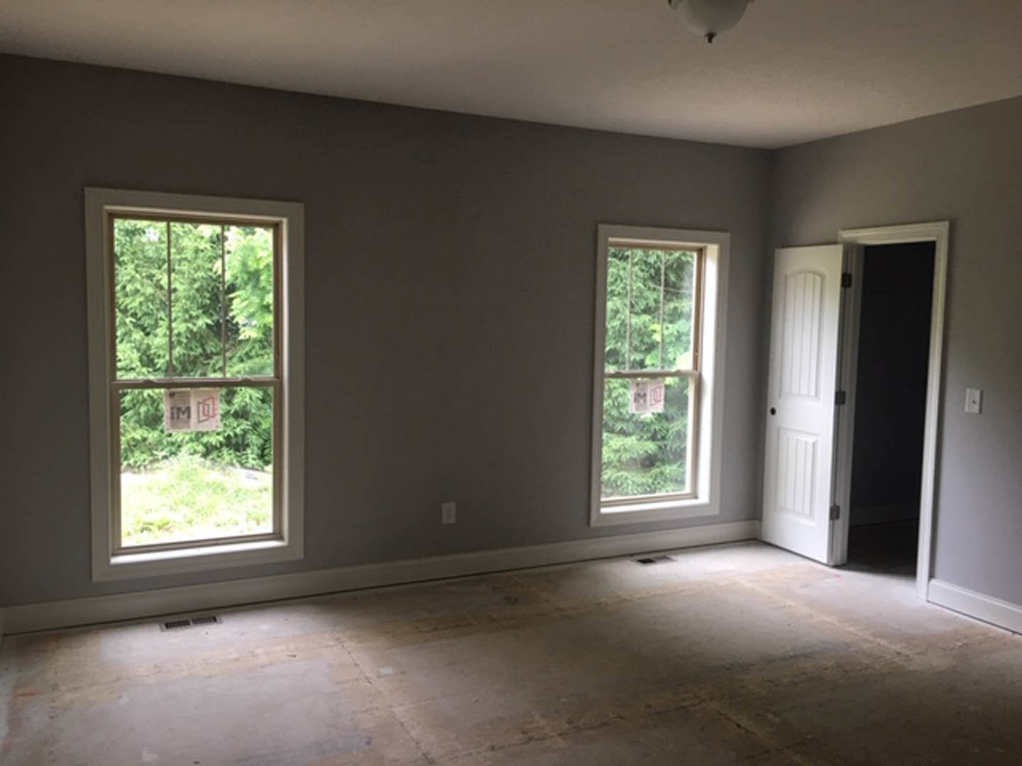 Bright room with two large windows, white door with black knobs, smooth concrete floor, and sunlight streaming across pale walls; one window displays a paper sign.