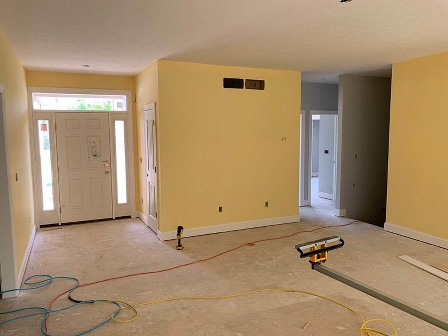 Yellow-painted walls, white door with glass panel and black handle, visible electrical cable on floor, wall with small holes, light-colored flooring