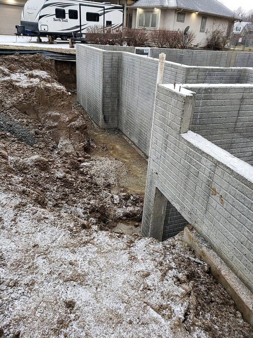 Concrete retaining wall with circular opening in snowy yard, white trailer parked nearby, house with satellite dish and illuminated window in background.