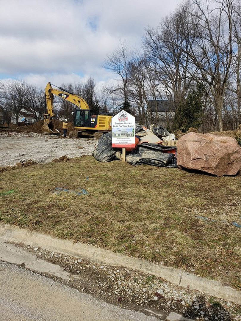 Construction site with grassy yard, large rock, leafless tree, crane-equipped vehicle, and close-up sign on the ground near paved road under cloudy sky