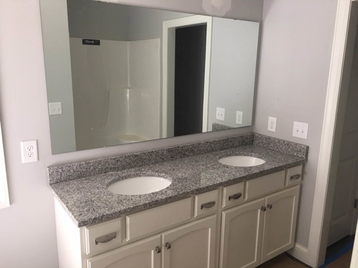 Modern bathroom featuring a rectangular mirror above a white countertop with integrated sink, chrome faucet, and minimalist cabinetry; visible electrical outlet and dual black-knob