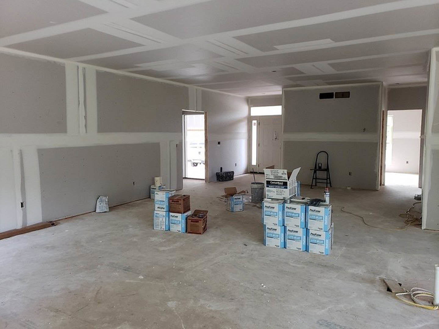 Unfinished room with plaster walls and ceiling, brown moving boxes scattered on the floor, black ladder holding a silver paint can, close-up of a white door, white object resting