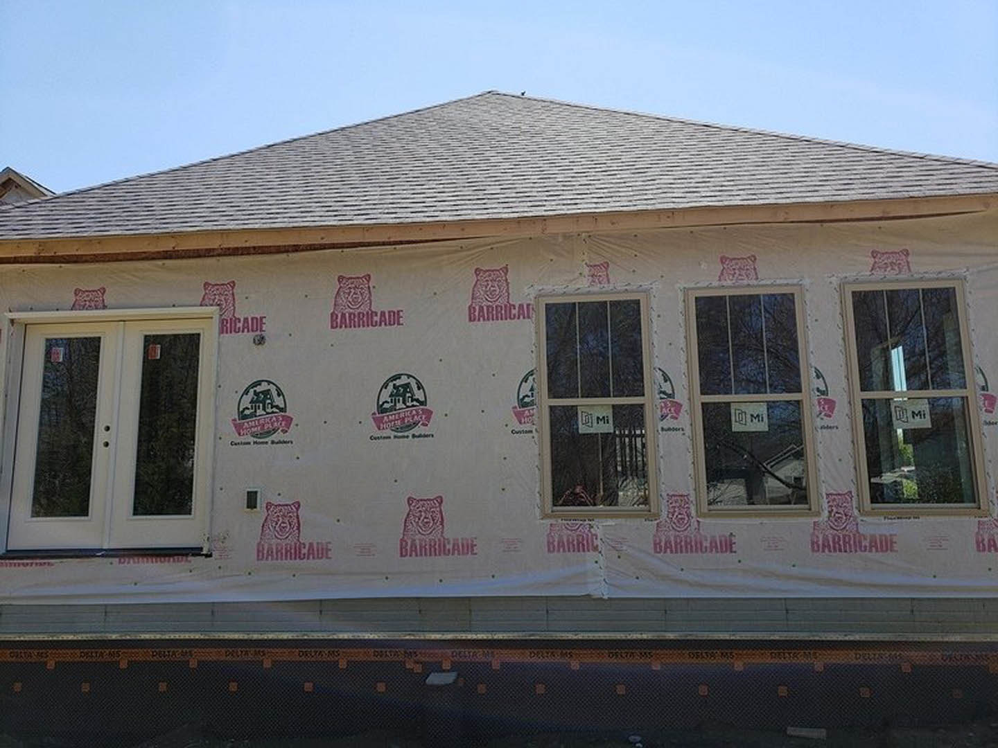 Partially built house with plastic sheeting covering the roof, double glass panel doors, and a window displaying a black and white sign with the letter M.