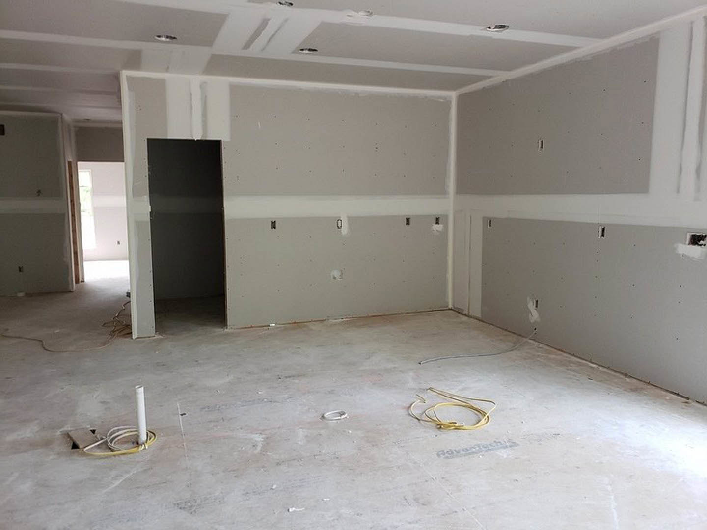 White-walled room with a black door framed in white, concrete floor scattered with white and yellow electrical wires, sunlight streaming across plaster walls, white ceiling