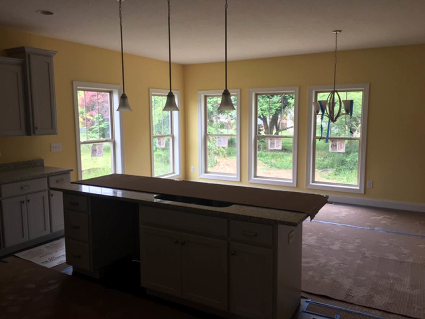 Modern kitchen with white countertops, stainless steel sink, brown flooring marked with blue tape, large windows featuring a sign and a white object, light cabinetry, and a