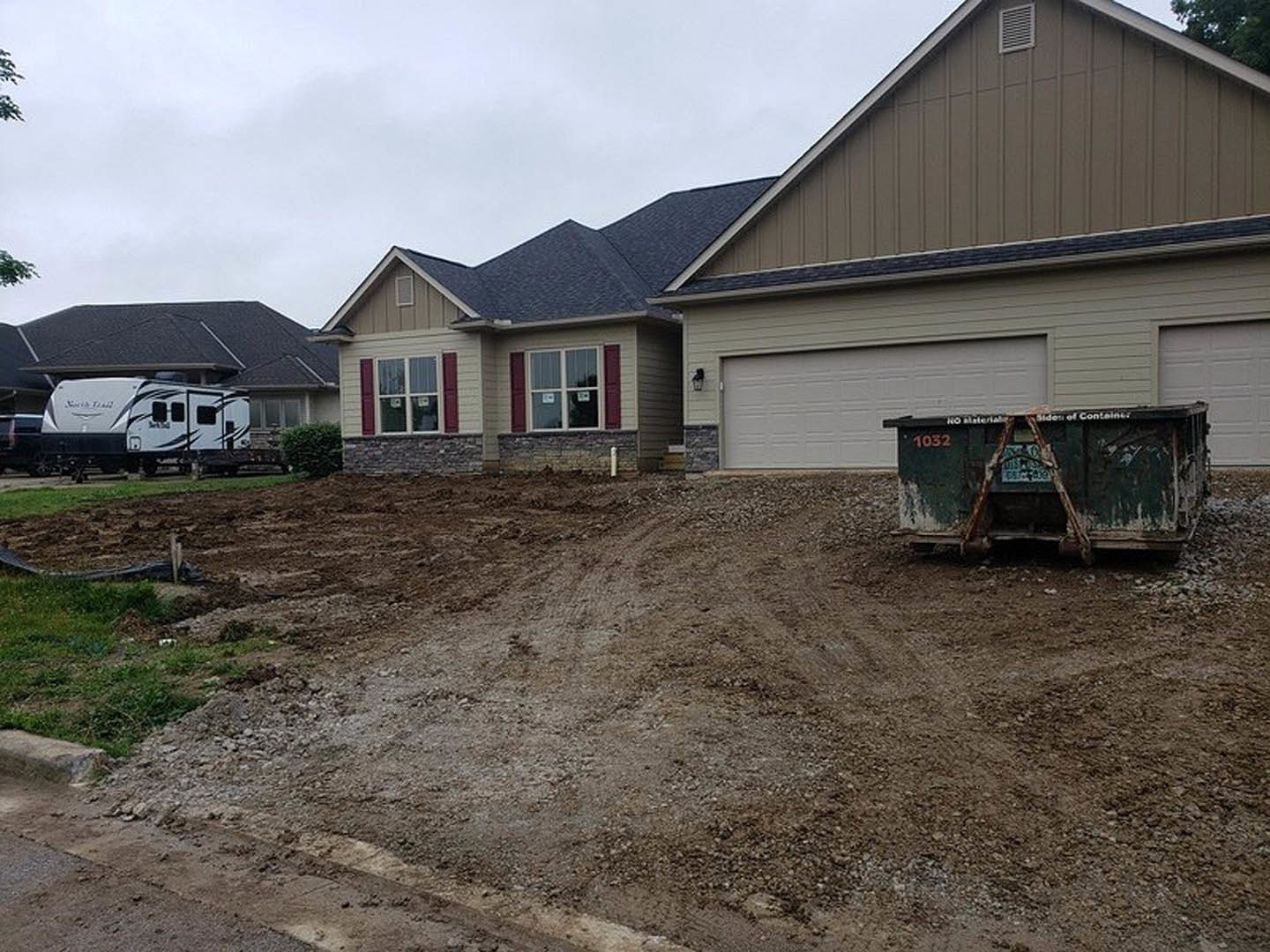 Dirt road leading to a house with a dumpster in front, white and black RV parked nearby, garage door visible, window with a sign, surrounded by trees and plants