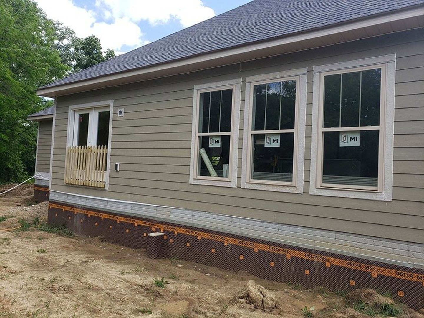 Two-story house under construction with exposed foundation, dirt yard, temporary fencing, and a window displaying a sign