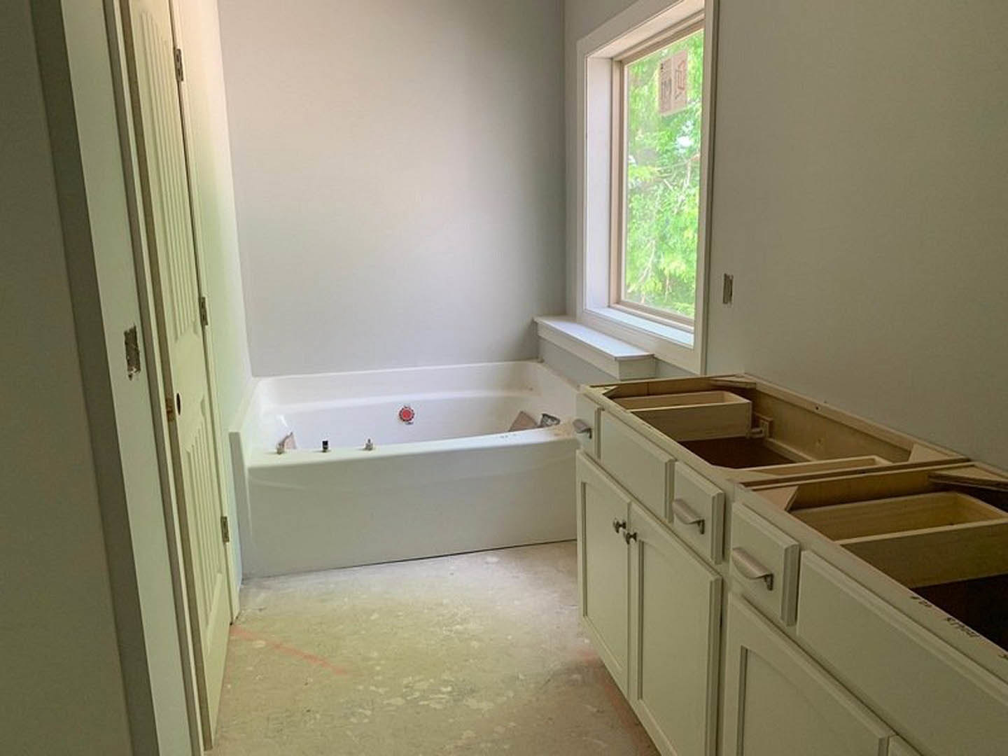 Bathroom with white bathtub featuring a red button, built-in sink, wood cabinets with drawers, large window overlooking outdoors, light-colored countertop, and tiled floor.