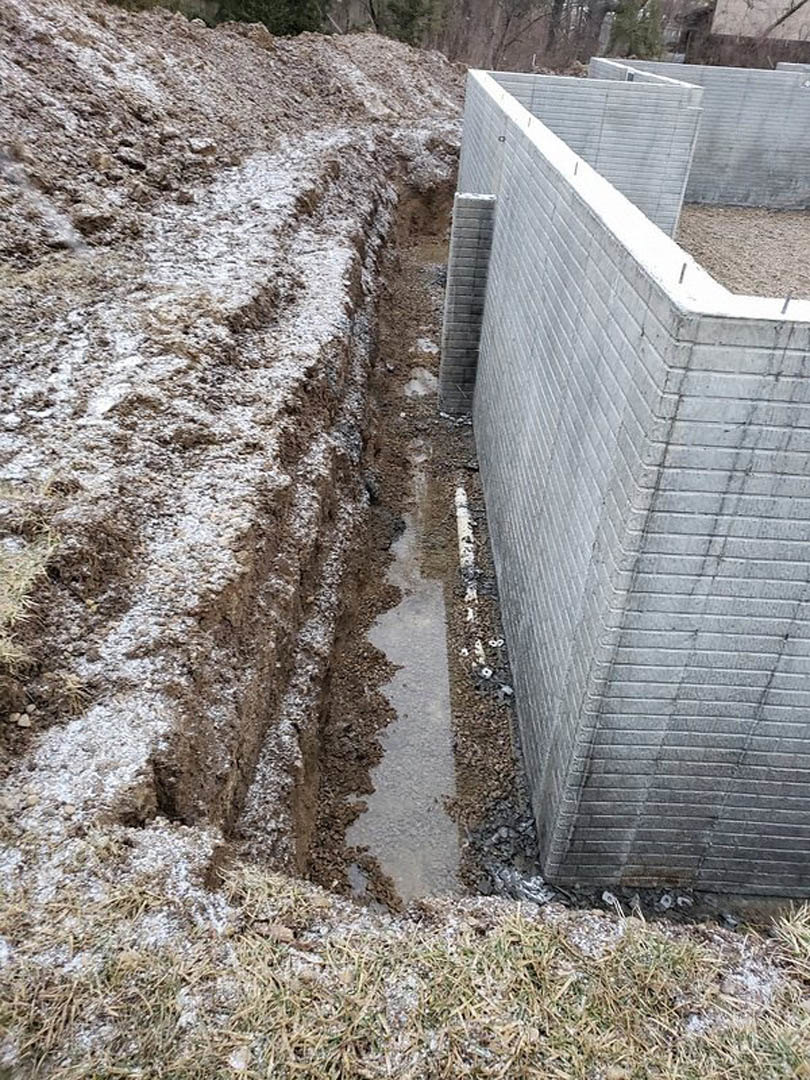 Concrete exterior wall with circular hole above a snow-dusted dirt trench, surrounded by grass and rocks