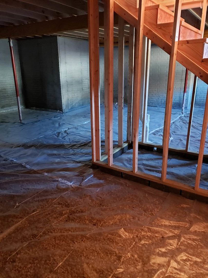 Unfinished basement with exposed wood framing, staircase, plastic bag on concrete floor, black wall featuring red handle, and visible dirt and sand surfaces