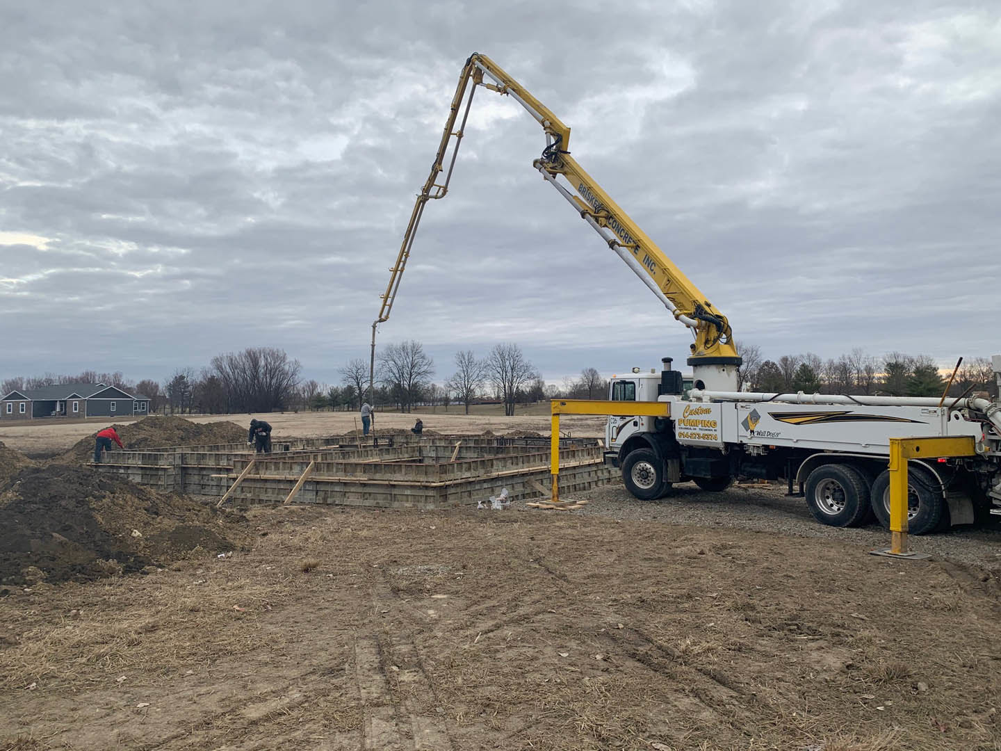 Yellow crane truck with concrete pump parked on dirt field beside custom home with grey roof; worker in red jacket stands near white truck with yellow and black text; cloudy sky