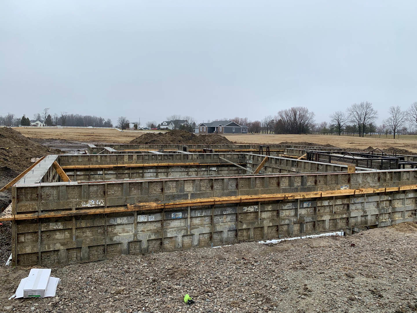 Concrete foundation with exposed wooden beams, surrounded by soil and rubble, under open sky with trees in background.