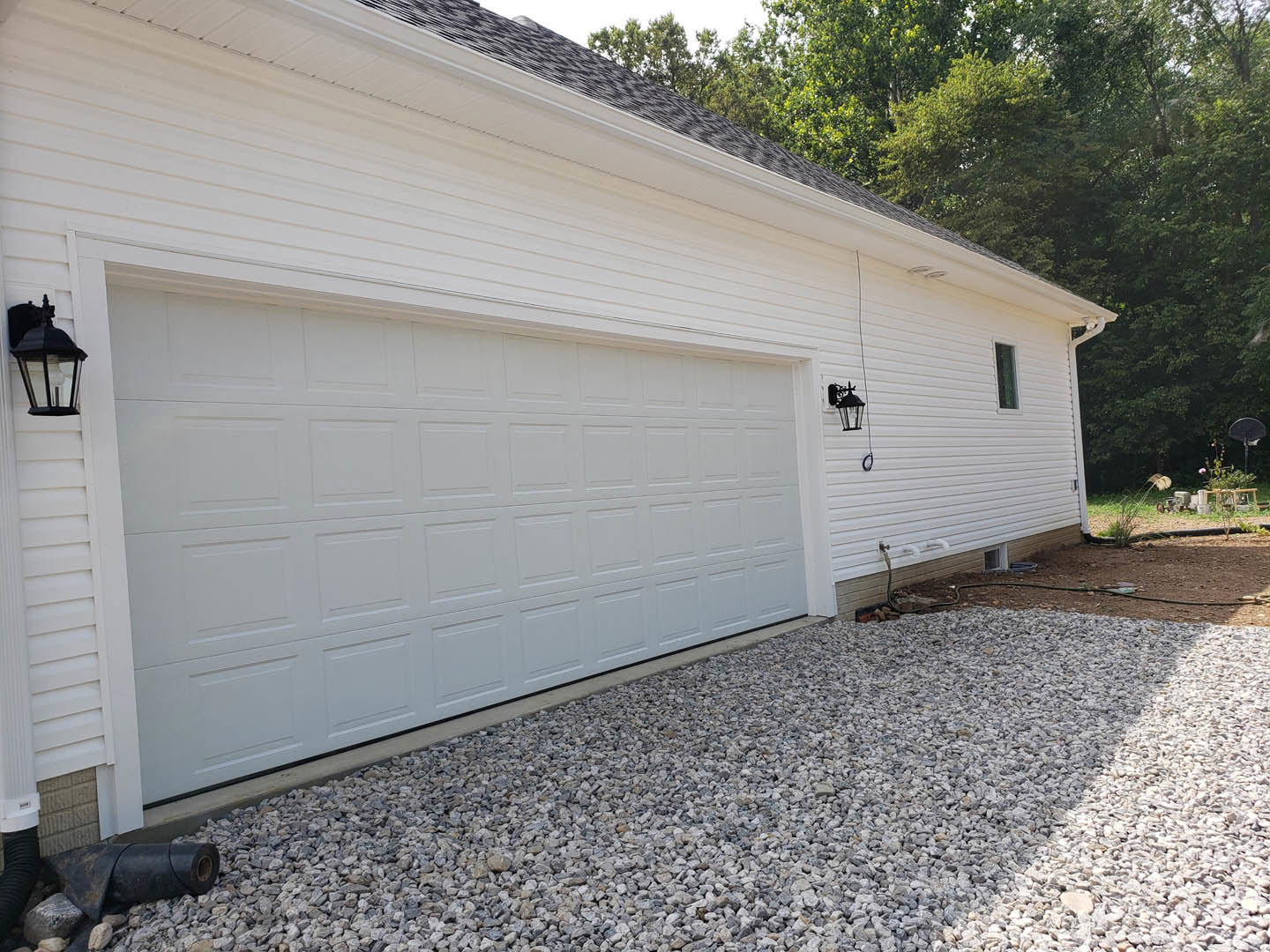 White garage door with horizontal panels, black cat perched near the center, white siding above, gravel driveway in foreground, black pipe lying on ground, exterior lamp mounted