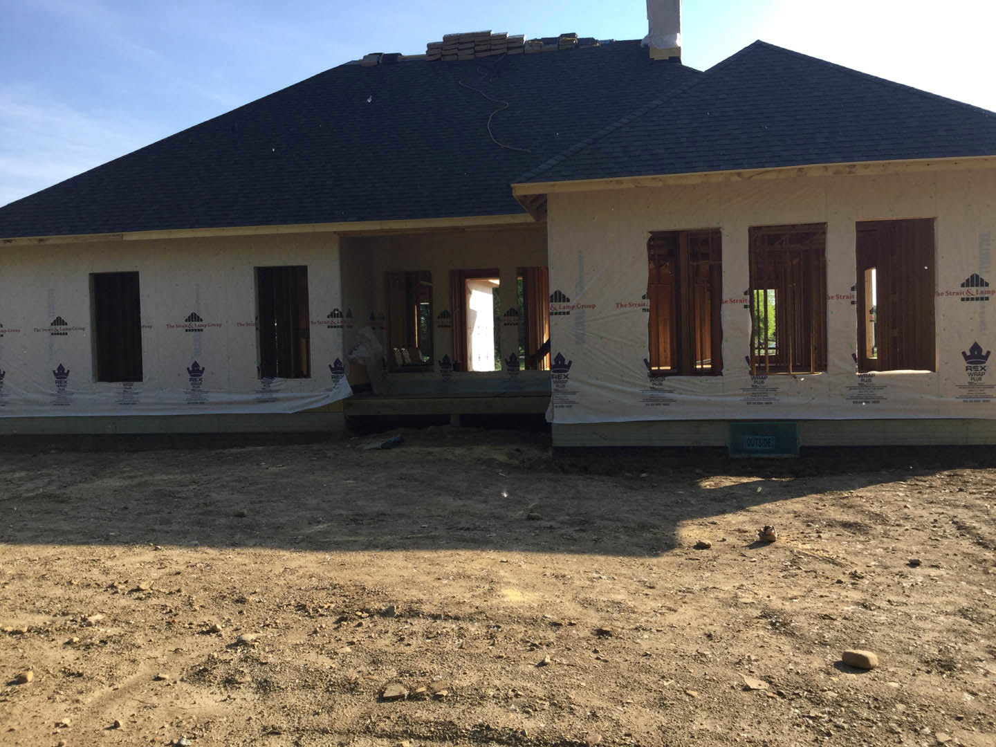 Partially built house with black roof, barred window, and unfinished dirt ground scattered with rocks