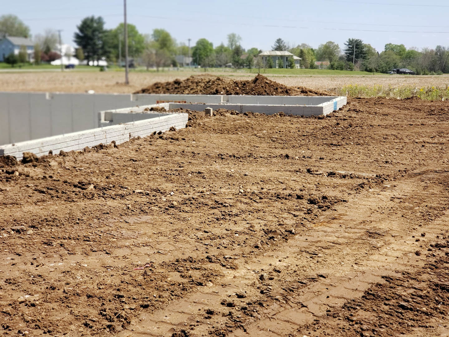 Dirt field with scattered soil and mud, foundation area visible, trees and grass in the background under open sky