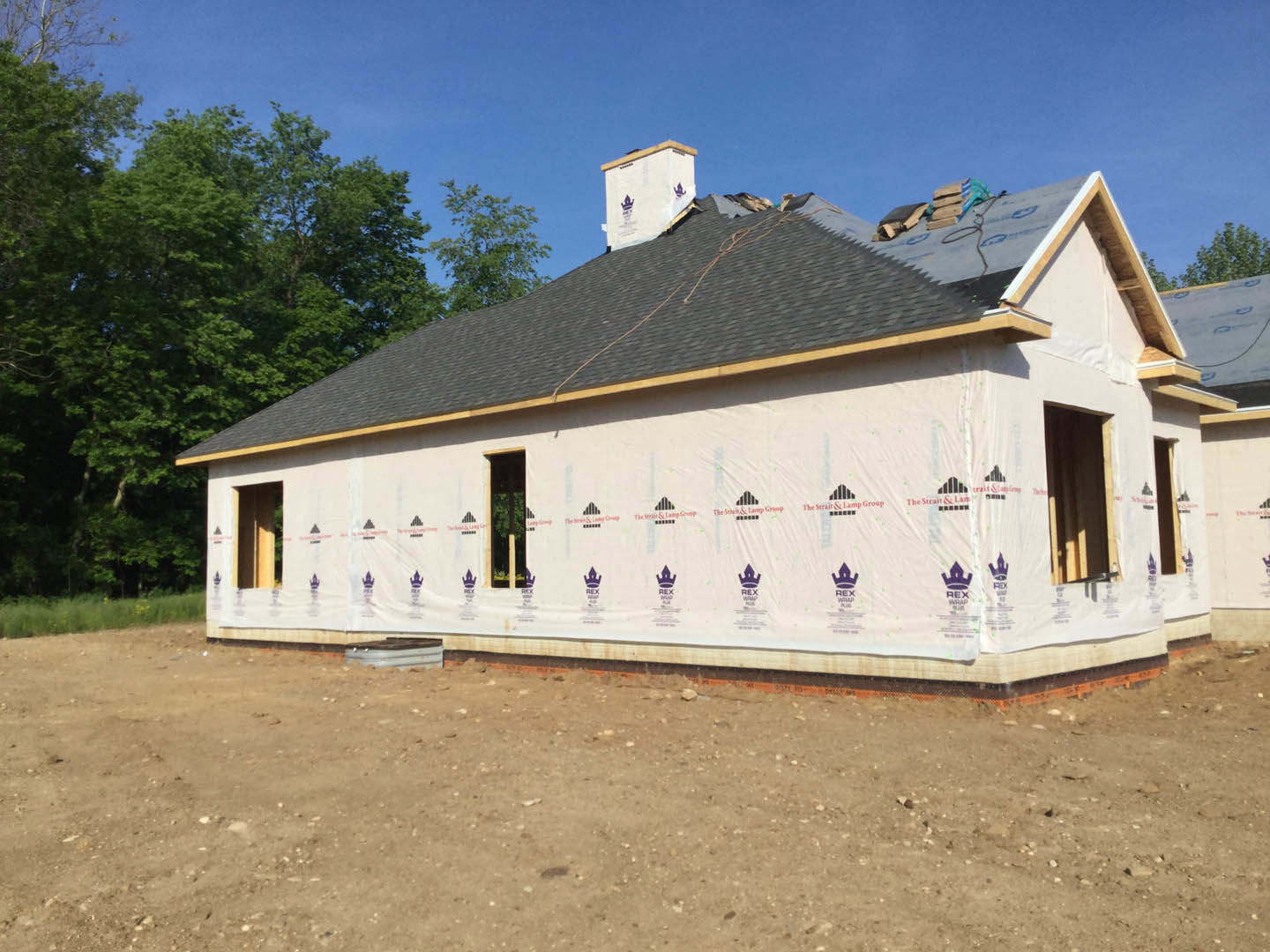 Framed house under construction with exposed roof trusses, dirt lot, white construction sign, and surrounding trees in the background
