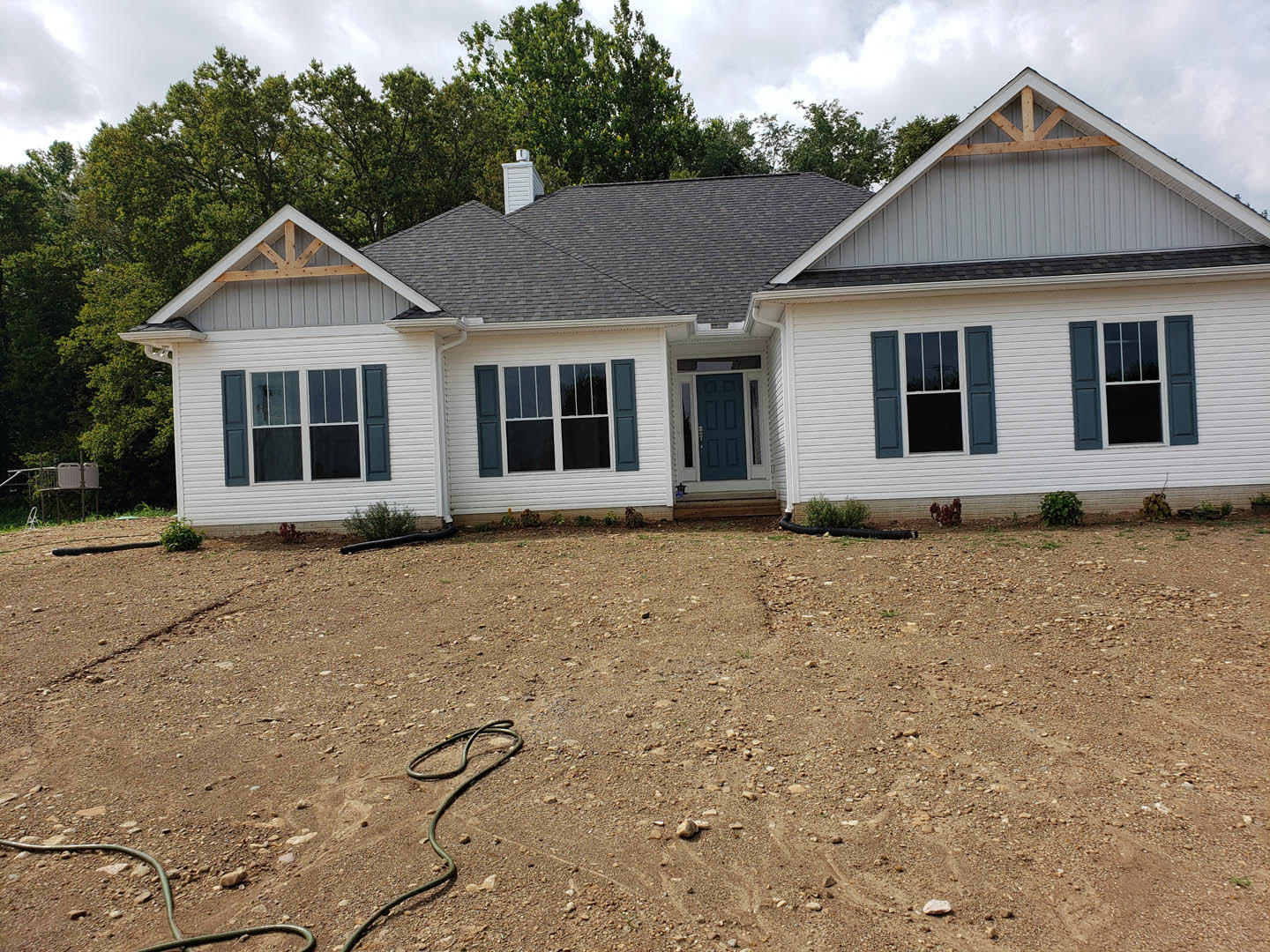 White cottage-style home with blue door and blue window shutters, surrounded by a dirt yard, garden hose lying on the ground, and a leafy tree nearby