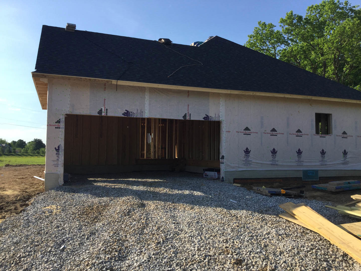 Framed house under construction with unfinished roof, large attached garage, piles of rocks and lumber on gravel driveway, surrounded by open land and trees