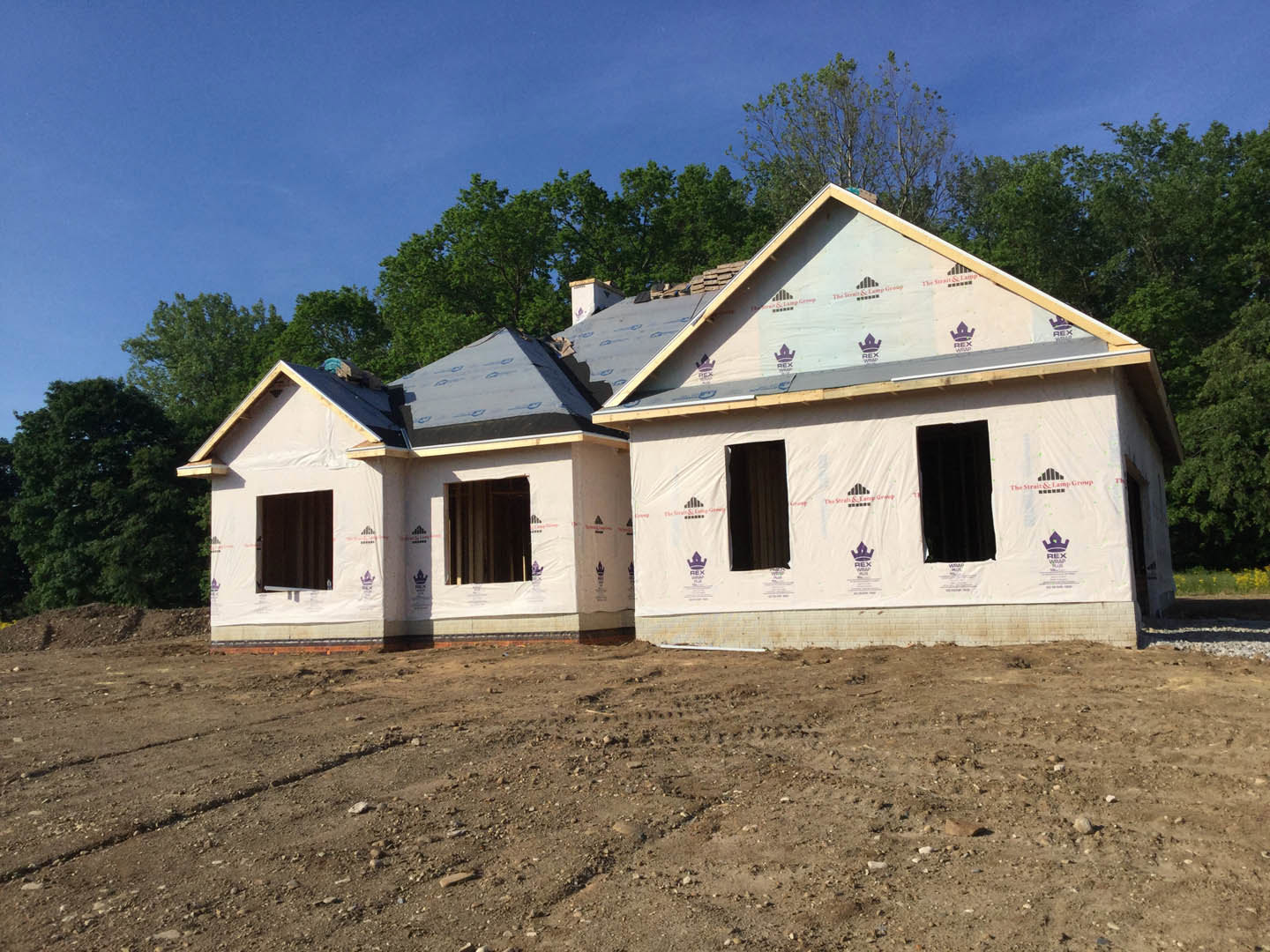 Wood-framed house under construction on dirt lot, surrounded by trees, partially installed windows, exposed roof trusses, cloudy sky overhead