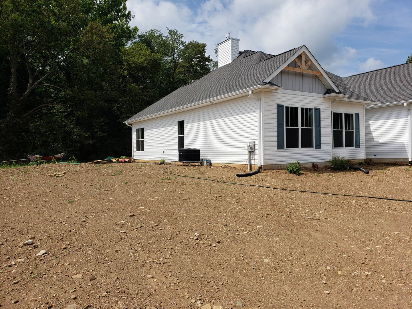 White cottage-style home with black roof, blue window shutters, dirt yard, scattered trees, garden hose, and bush in foreground.
