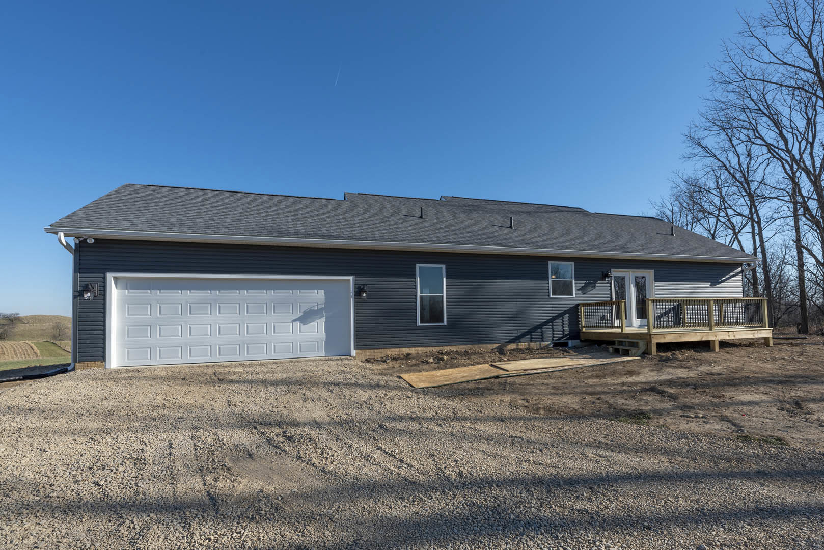 Two-story house with white-framed windows, attached garage with paneled door, gravel and wood driveway, wooden deck featuring metal railing, surrounded by trees and open sky