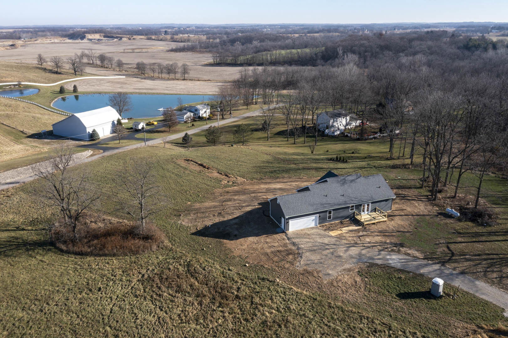 Two-story white house with gray roof, attached garage, wooden deck with railings, surrounded by grassy field, lake in background, mature trees nearby