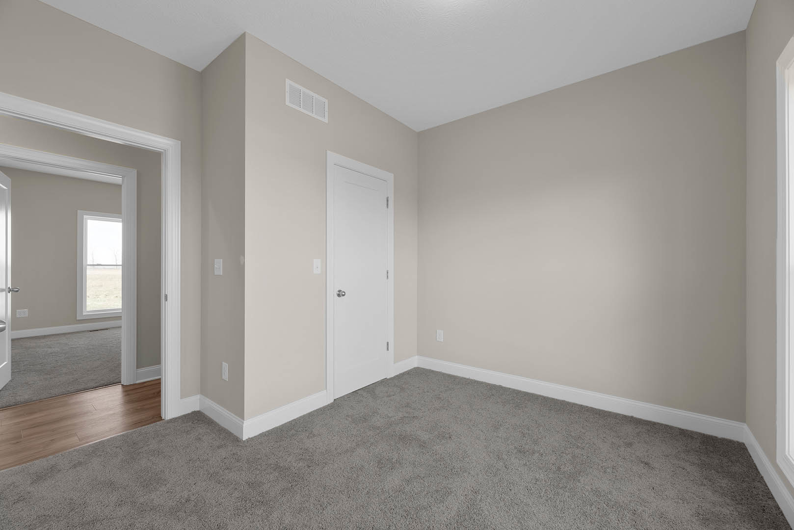 White door with silver knob set in a white door frame, grey carpet flooring, hallway with wood floor, window overlooking grassy field, light-colored walls with molding.