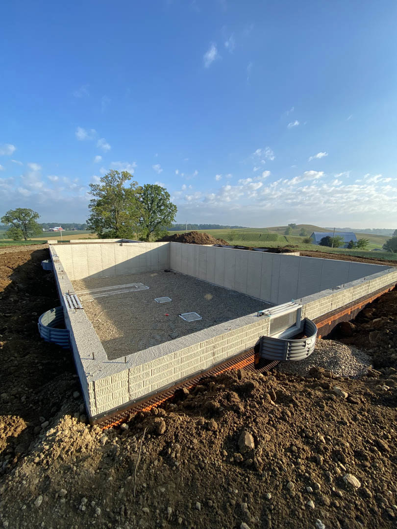 Concrete foundation walls and footings surrounded by piles of dirt, construction materials, and grassy field with trees under a partly cloudy blue sky