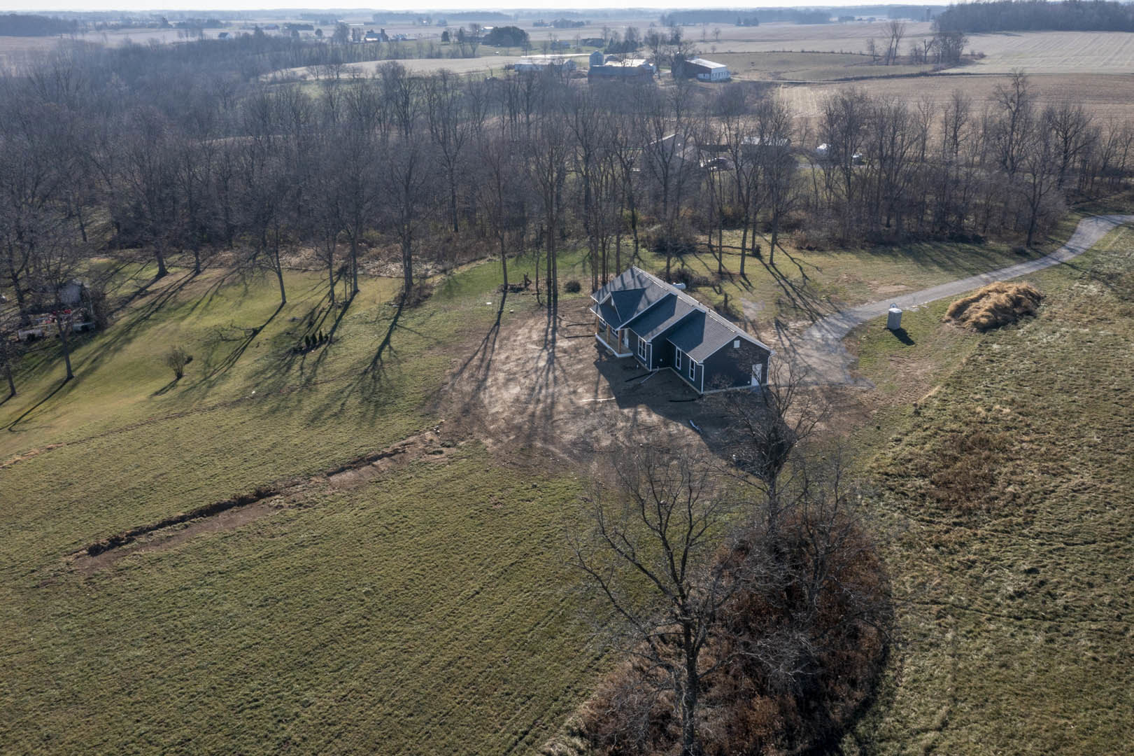 White custom home with dark roof sits in open grassy field, surrounded by leafless trees and wooden fence, under pale winter sky