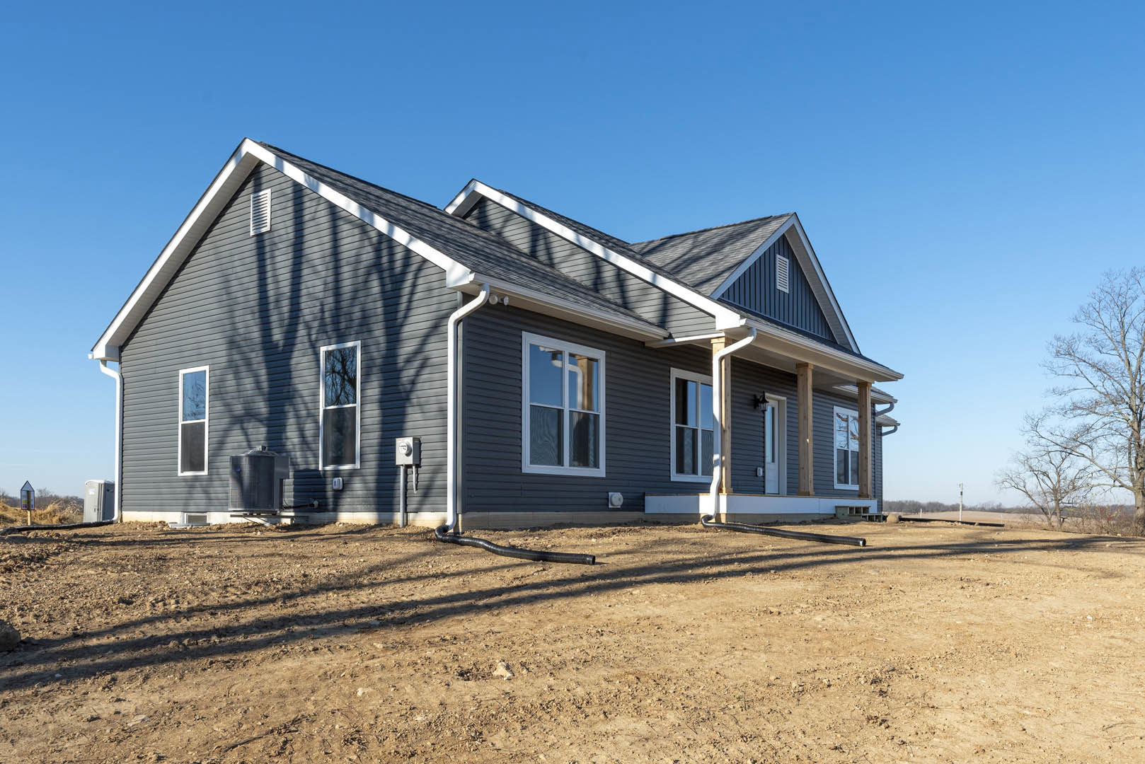 Partially built house with white roof, exposed framing, white window frames, bare tree nearby, black pipe lying on dirt under clear blue sky