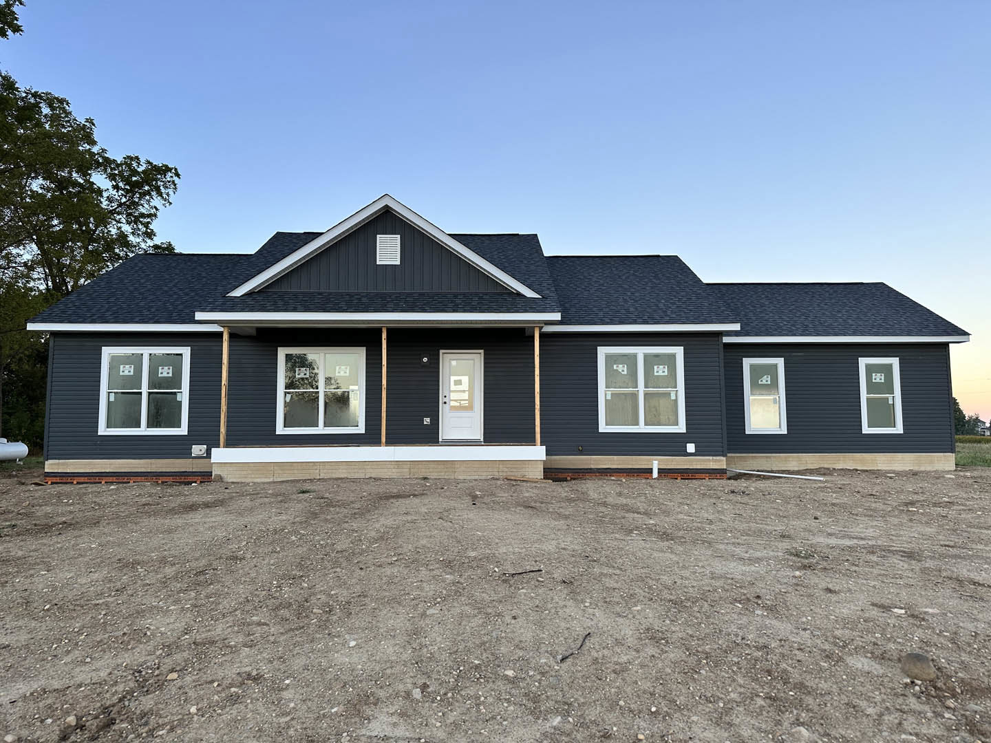 White-framed windows and door on a partially constructed house with exposed dirt ground, light siding, and visible tree in background