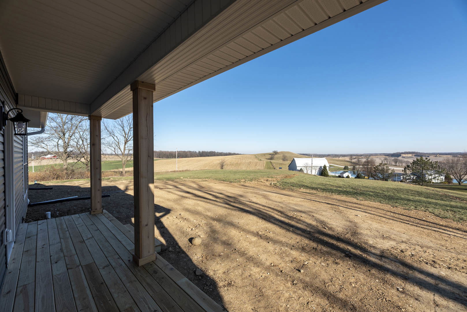 Wooden porch with white pillars overlooking a dirt field, distant white house surrounded by trees, grassy areas, and clear sky