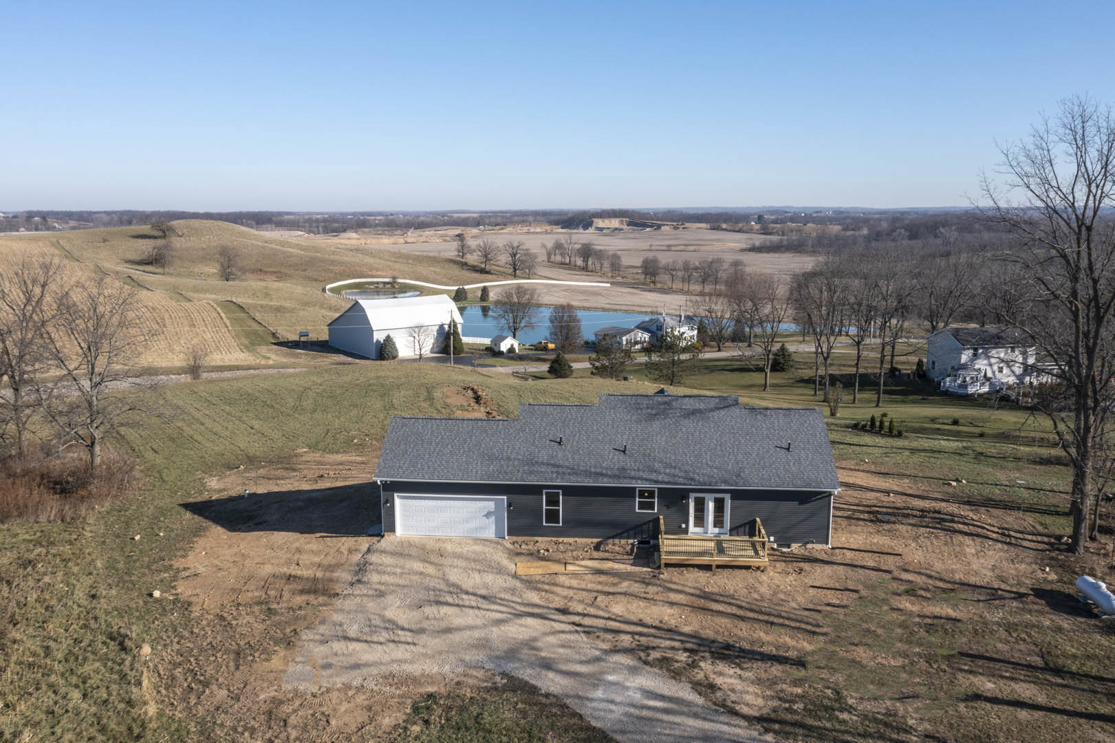 White farmhouse with black trim and deck railing, surrounded by grassy field, trees, and pond under blue sky with clouds