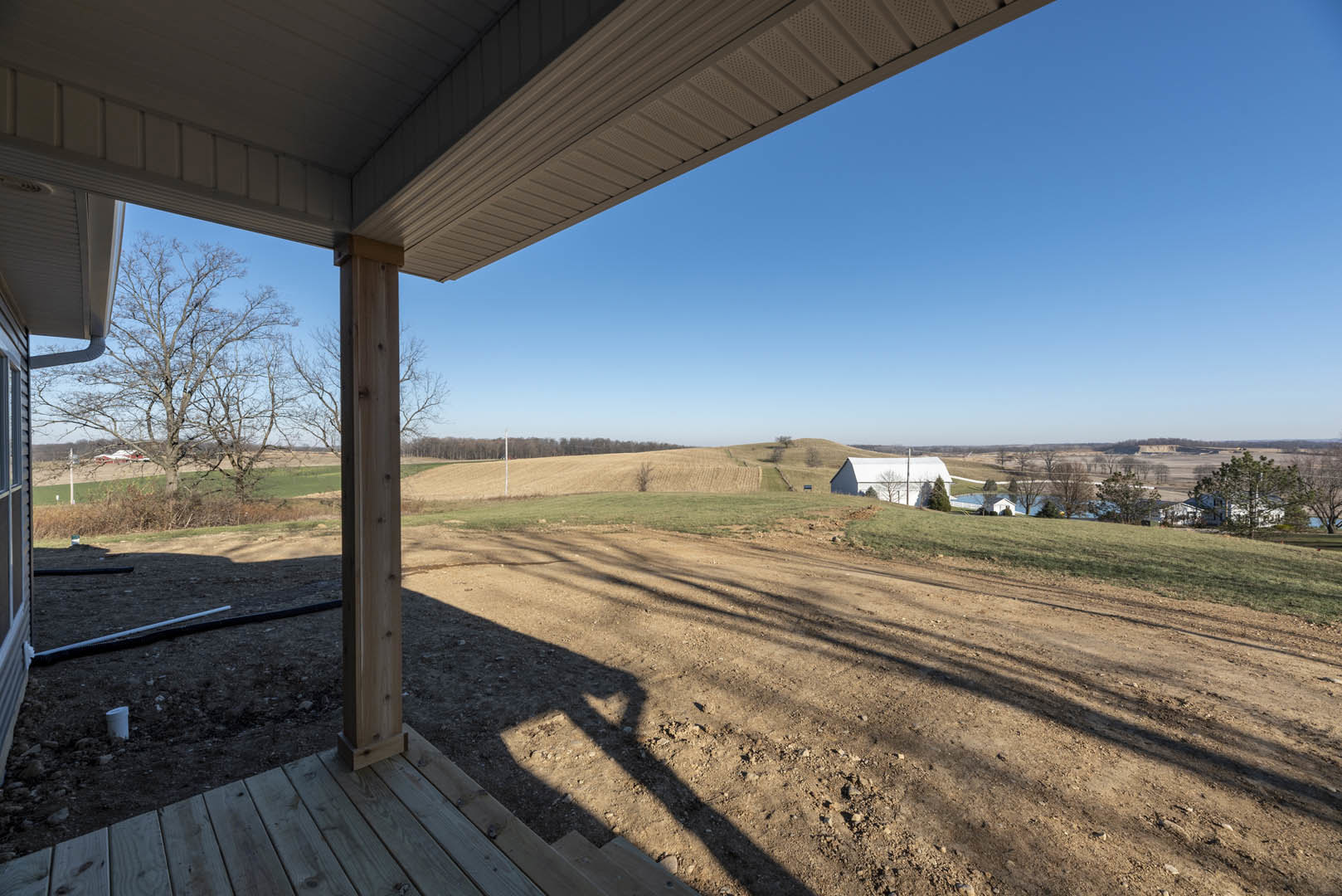 Wooden porch with vertical post overlooking dirt field, shadow cast on ground, person standing near unfinished land, house with gable roof in background, trees and sky visible