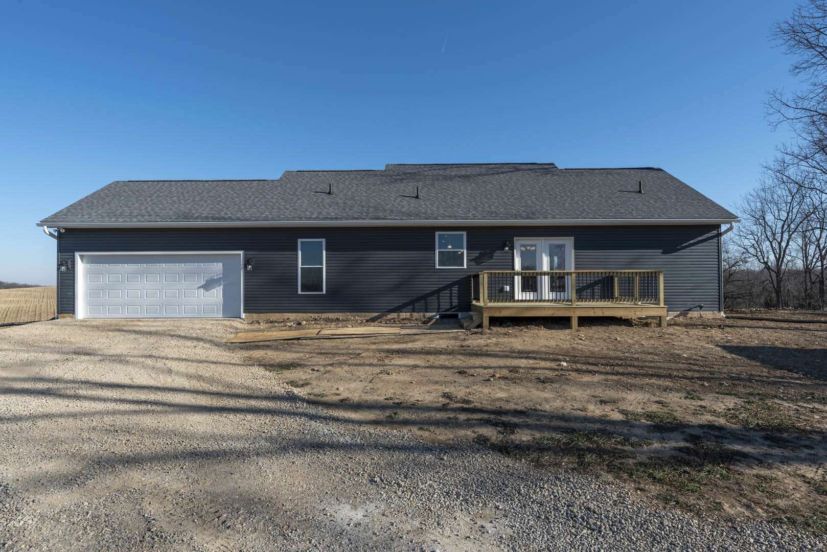 Wooden deck with railing overlooking dirt yard, white garage door beneath black roof, blue roof accents, white-framed windows, light-colored siding, surrounded by trees and open