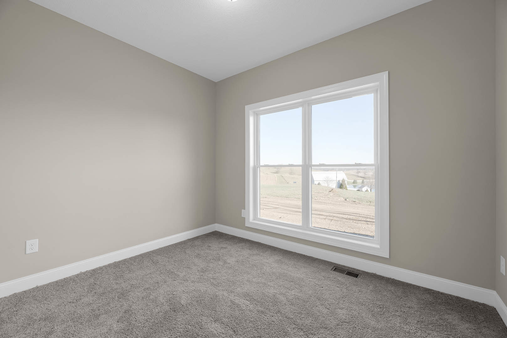 Neutral-toned carpeted room with white walls, large window overlooking farmland, white ceiling, and visible electrical outlet