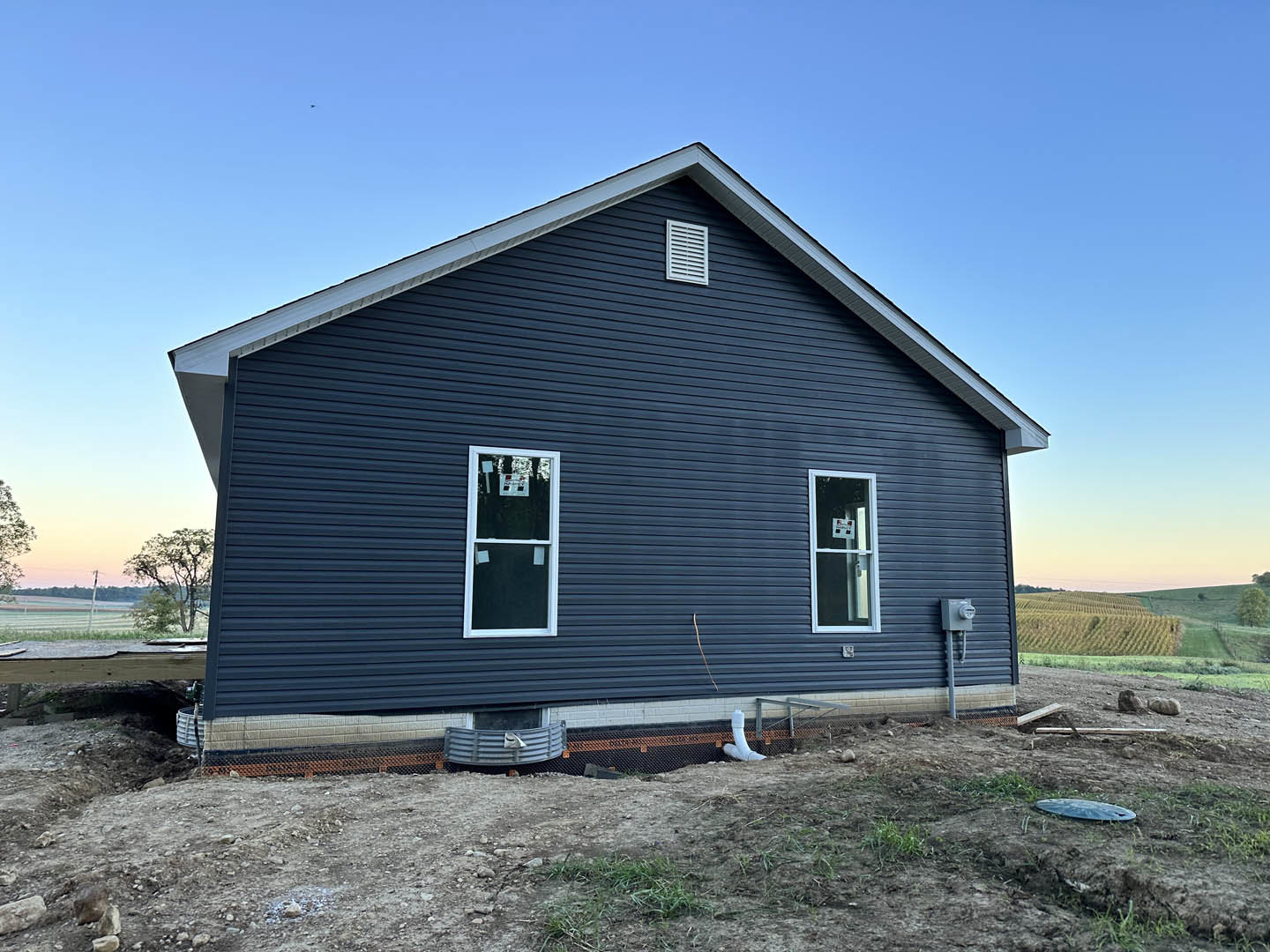 Two-story house under construction with blue siding, white-framed windows, blue roof, white vent, and construction sign on window, set against clear blue sky and grassy yard
