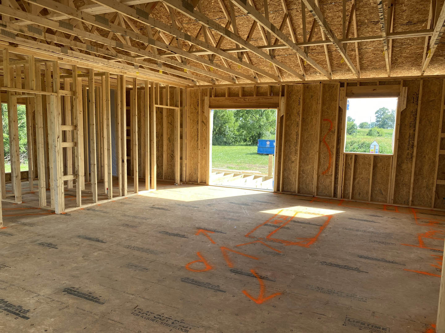 Concrete floor marked with orange paint and arrows, exposed wood roof truss overhead, large window framing view of grassy field and trees, blue dumpster visible outside