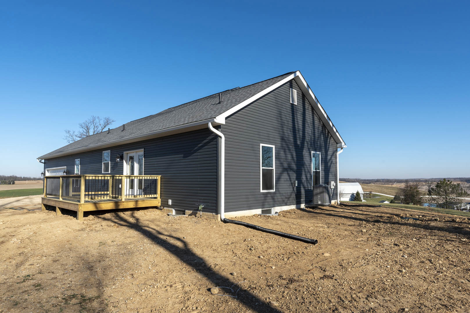 Two-story house with white-framed windows, wooden deck featuring black railing, fenced yard with grass and dirt, black pipe near ground, blue sky overhead, trees in background